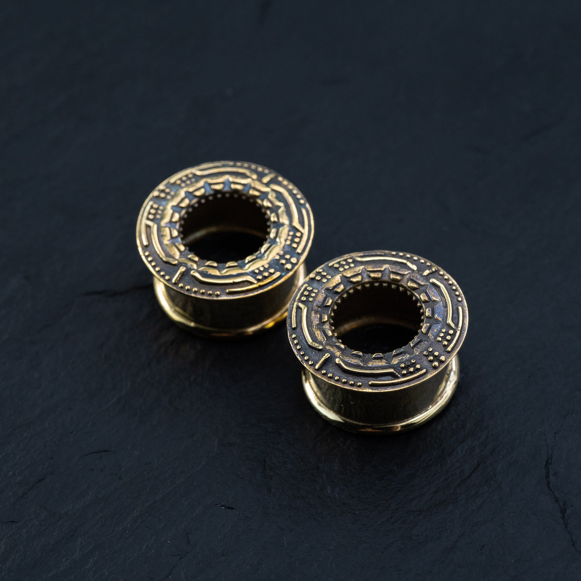 Close-up of two Kaya Movement TERA Futuristic Sci Fi Flesh Tunnels in gold brass on a dark surface. The central opening and engraved details are prominent, giving them a mechanical and ornate appearance reminiscent of futuristic ear tunnels. Available in sizes ranging from 8mm to 20mm.